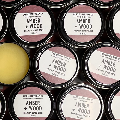 Multiple small tins with rose colored labels indicating the scent 'Amber and Wood' beard balm. One of the tins has the lid off showing the balm inside.