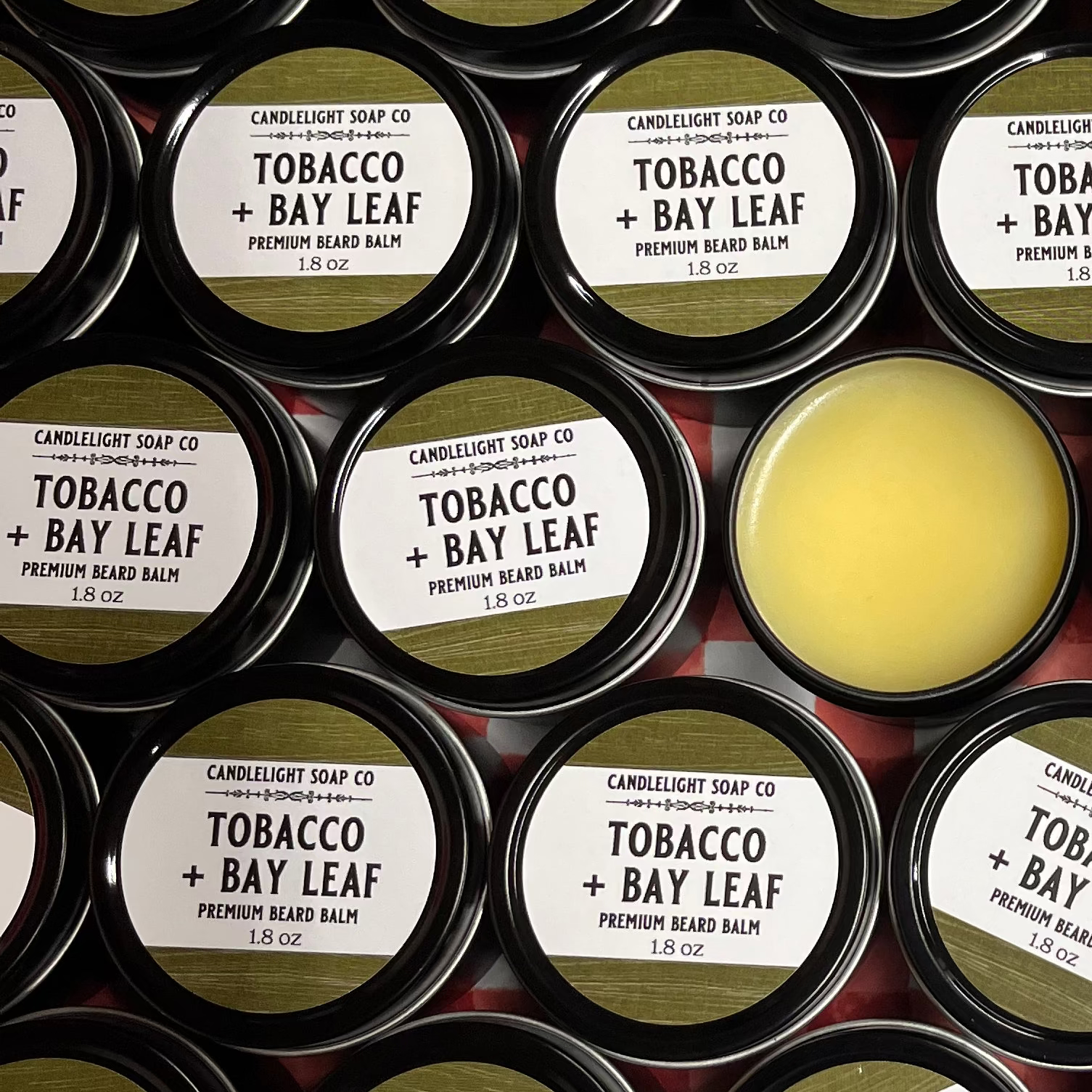 Collection of small tins with green labels indicating the scent 'tobacco and bay leaf'. One of the tins has the lid off to show the balm inside.